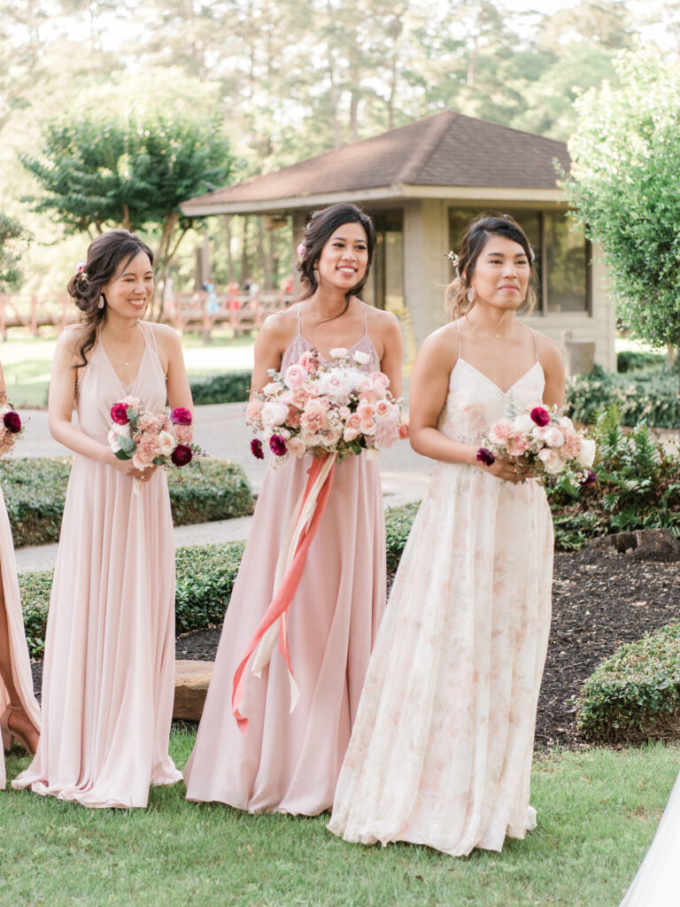 Bridesmaids wearing shades of blush, look on smiling as their friend exchanges vows with her groom. 