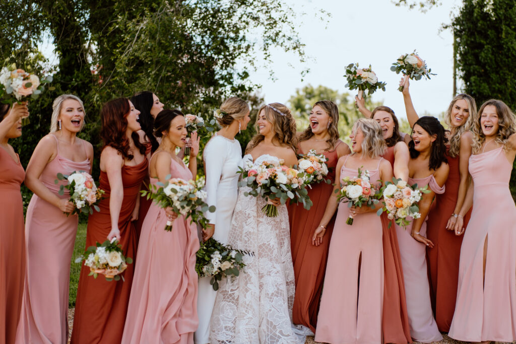 Two brides lean in for a kiss as their bridesmaids, in various shares of pink, peach, and rust, cheer them on.