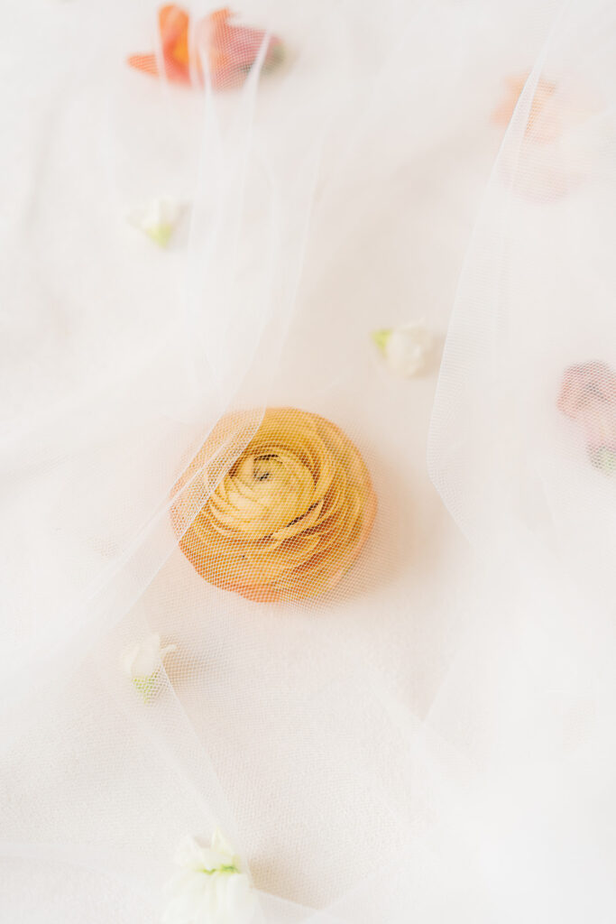 A veil lays over a scattering of flowers including small white buds, peach snapdragon blooms, and a soft orange ranunculus.