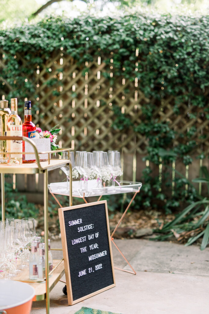 A gold and wood bar cart is topped with liqueurs for sprtiz drinks, an acrylic side table holds wine glasses with baby's breath florals tied to the stem, and a letterboard sits in front reading "Summer Solstice: Longest Day of the Year; Midsummer. June 21, 2022"  